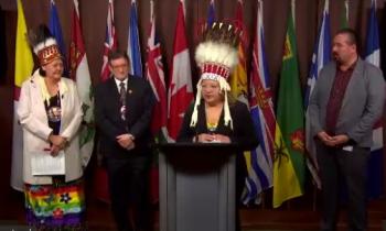 A woman in a chiefs headdress stands at a podium surrounded by other chiefs.