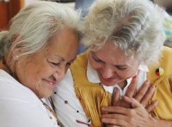 Two older woman share a time together. One has brought the others hand and placed it over her heart. They bring their heads together with affection.