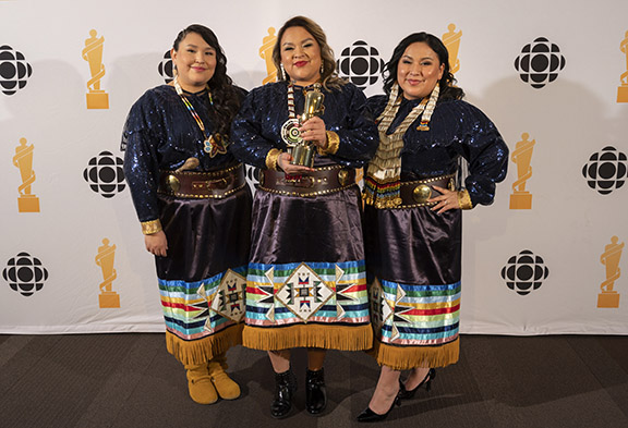 Three women in ribbon skirts. The one in the middle hold a JUNO award..
