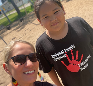 A woman and boy are photographed. They are smiling. He wears a black t-shirt that reads National Family Violence Prevention Month, with a red painted hand print on it.