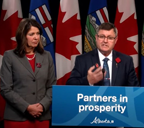 A woman stands beside a man who is at a podium. The woman is Premier Danielle Smith and the man speaking into a microphone is Minister Rick Wilson.