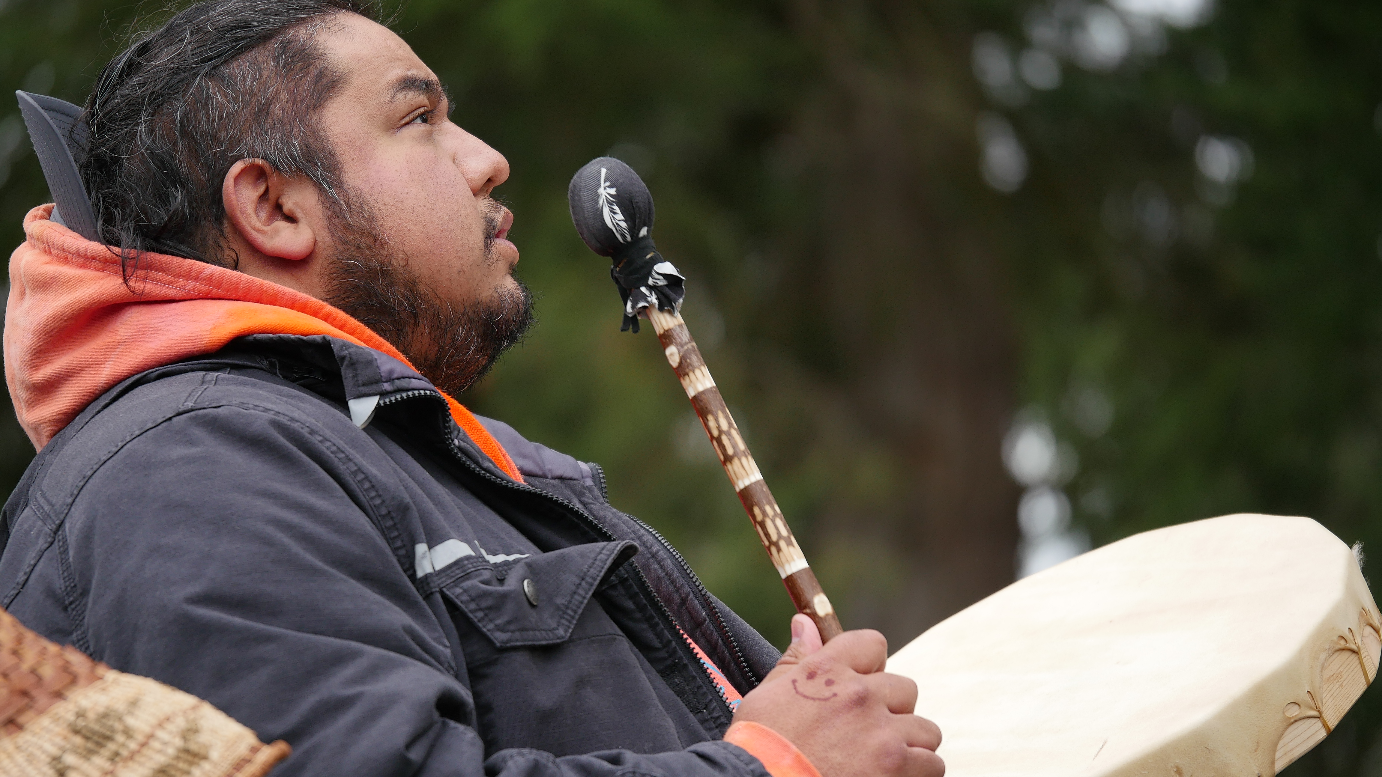 A man looks to the skies as he plays a hand drum. He is standing outdoors.