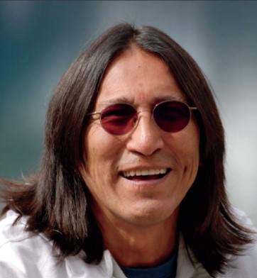 A head and shoulders photo of a man wearing dark glasses. His hair is parted in the middle and hangs to his shoulders. He is smiling.