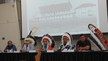 Chiefs in headdresses sit in front of a photo of a residential school