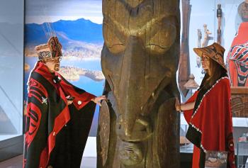 A man and a women dressed in button blankets and cedar hats stand with their hands on the totem pole.