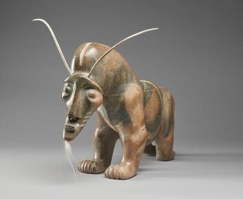 A mottled brown soapstone carving shows a muscular Muskox with an exaggerated animated face with a tuft of white fur coming out of his chin and narrow horns sprouting from the top of its head. 
