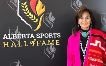 A woman stands against a grey wall with logos printed on it and the words Alberta Sports Hall of Fame. She wears a bright pink jacket and has a folded red blanket draped over her shoulder.