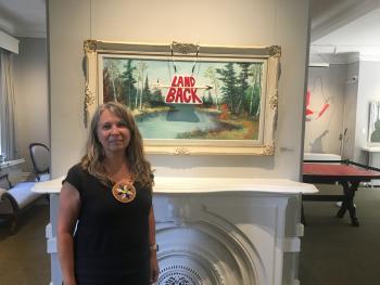 A woman stands in from of a framed painted landscape. Hanging in the middle of the painting is a beaded medallion that says "Land Back"