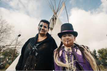 A woman and boy stand outdoors in front of a tipi. Their faces are painted with black and white paint. He wears a top hot with a purple ribbon wrapped around it. He has on a purple vest with two white feathers appliqued on the shoulders.