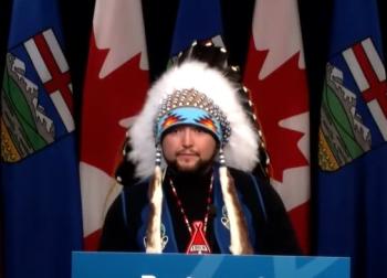 A man standing at a podium wears a chief's feathered headdress as he speaks into microphone.