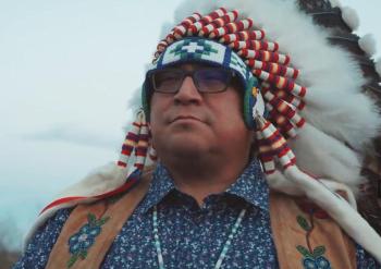 A man wearing a chief's feather bonnet looks off into the distance.