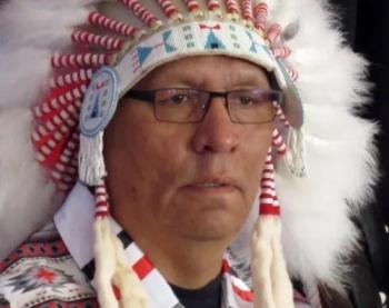 A close-up of the face of a man wearing a feather chief's bonnet is seen.