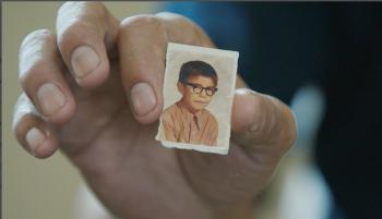 A small school photo of a boy wearing dark-rimmed glasses is held up between the index finger and thumb of a man's hand.