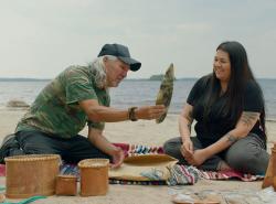 Two people sit on a beach with a body of water behind them. They are surrounded by a display of birchbark baskets, and the older man is holding some artifact for the younger woman to look at.