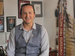 A smiling man wearing a shirt with a suit vest over top sits perched on the edge of a desk. On the walls behind are photographs of graduates and diplomas. Behind his is the eagle staff of the Cowesses First Nations.