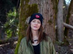 A young woman stands against the backdrop of old growth trees. Moss is growing up one side of a big tree directly behind her. She wears a black toque with an N in white on a red circle on the hat.