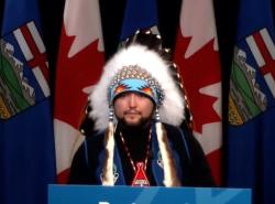 A man standing at a podium wears a chief's feathered headdress as he speaks into microphone.