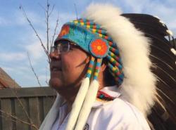 A man stands outside in a yard. He wears a chiefs feather headdress.