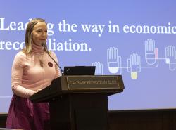 A woman stands at a podium in front of a screen with the words "Leading the way in economic reconciliation" printed on it.