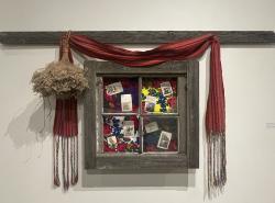 An old house board and window hangs on a gallery wall. Inside the frames of the window are photographs. A Métis sash is draped above the window an a bouquet of dried flowers hang upside down on the left side of the window frame.