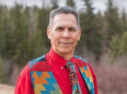 A man in a red shirt and multi-coloured vest and tie stands in front of a backdrop of trees.